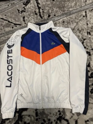 Lacoste vindjacka blå orange vit - Snygg vindjacka från Lacoste i vitt med blå och orange panel framtill samt svarta detaljer. Jackan har dragkedja, hög krage och resår i midja och ärmslut. Stor Lacoste-logga på ena ärmen och klassisk krokodil på bröstet. Perfekt för en sportig streetwear-look.