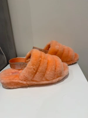 Fluffiga orange tofflor från UGG - Säljer ett par supermjuka och fluffiga tofflor från UGG i en härlig orange färg. Tofflorna har en bred rem med glittrande detaljer och öppen tå. Perfekta för att mysa runt hemma i och ger en lyxig känsla till vardagen.