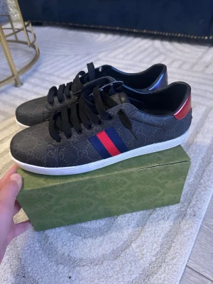  Gucci ace svart  - Snygga svarta Gucci sneakers med klassiskt GG-mönster, vita sulor och svarta skosnören. Sidan har en röd och blå rand och hälen har en röd respektive blå detalj. Tillverkade i läder med textilinslag. Perfekta för dig som vill ha en lyxig och stilren look det är fejk men ingen box inkluderad 