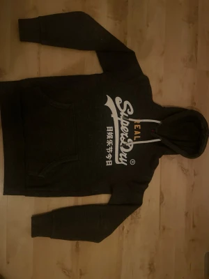 Mörkgrå Superdry hoodie med tryck - Mörkgrå hoodie från Superdry med stor vit logga och japansk text på bröstet. Klassisk känguruficka framtill och justerbar huva med dragsko. Tillverkad i mjukt bomullsmaterial, perfekt för en avslappnad streetwear-look. Storlek S men kan fungera bra för M.