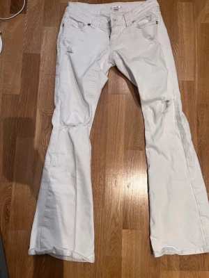 Vita bootcut jeans med slitningar - Säljer ett par vita bootcut jeans med slitna detaljer på låren och knäna. Jeansen har fem fickor, låg midja och utsvängda ben. Perfekta för dig som gillar en avslappnad och trendig stil. Materialet är bomull med lite stretch för extra komfort.