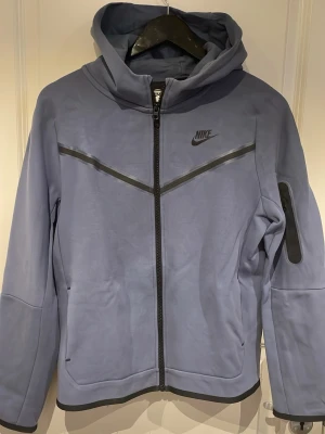 Blå Nike tech fleece hoodie med dragkedja - Snygg blå hoodie från Nike med hel dragkedja framtill och svarta detaljer. Hoodien har en huva, två sidofickor och en extra ficka med dragkedja på ärmen. Tillverkad i mjukt material som känns bekvämt och sportigt. Perfekt för en avslappnad streetwear-look.
