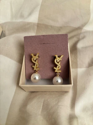 YSL örhängen med pärldetalj - Snygga örhängen i guldtonad metall med glittrande stenar och hängande vita pärlor. Logotypen YSL är tydligt synlig och ger en lyxig känsla.