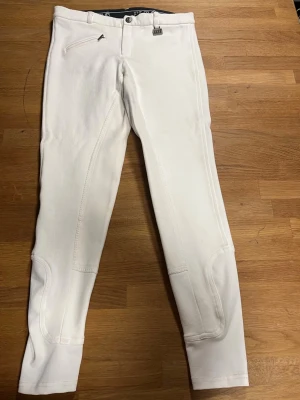 Vita ridbyxor från ELT - Snygga vita ridbyxor från ELT med förstärkta knäpartier och en dragkedjeförsedd ficka framtill. Byxorna har bälteshällor. Perfekta för träning eller tävling inom ridsport. Barn