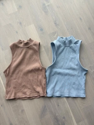 Ribbad ärmlös topp  - Två ribbade ärmlösa toppar , en i ljusblått och en i beige. Båda har hög hals och croppad passform, perfekta för en trendig och avslappnad stil. Materialet är mjukt och stretchigt, vilket gör dem bekväma att bära hela dagen.