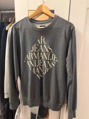 Grå långärmad topp Armani Jeans - Snygg grå långärmad topp från Armani Jeans med stort tryck framtill i vitt. Klassisk rund halsringning och normal passform. Perfekt för dig som gillar stilrena och trendiga plagg med logga.  PASSAR UNGEFÄR M/L