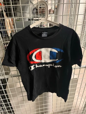 Svart Champion t-shirt med logga - Svart t-shirt från Champion med stor färgglad logga i rött, vitt och blått på bröstet. Klassisk rund halsringning och korta ärmar. Tillverkad i mjuk bomull som känns skön mot huden. Perfekt för en avslappnad och sportig stil.