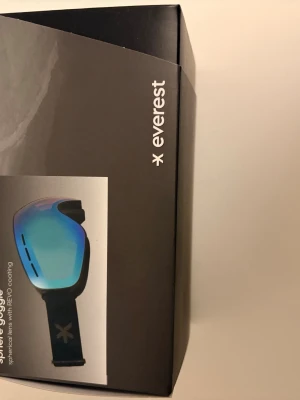 Blå Everest skidglasögon - Säljer ett par snygga skidglasögon från Everest med blå spegellins och svart ram. Glasögonen har justerbart band med Everest-logga och sfärisk lins med REVO-coating för extra skydd mot sol och snö. Perfekta för vintersport och skidåkning.