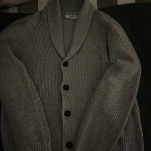Grå kofta från Lindbergh - Säljer en stilren grå kofta från Lindbergh i storlek S. Koftan har en avslappnad passform med ribbade detaljer och knappar framtill. Perfekt för kyligare dagar.