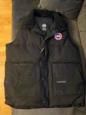 Svart dunväst från Canada Goose - Säljer en svart dunväst från Canada Goose med klassisk logga på bröstet och två stora fickor framtill. Västen har hög krage, tryckknappar och dragkedja. Perfekt för lager-på-lager och håller dig varm under kyliga dagar.