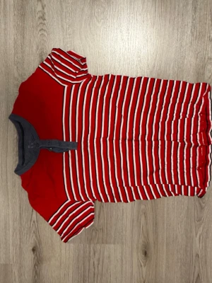 Röd och vit randig t-shirt - Snygg t-shirt med röda och vita ränder samt grå knappslå vid halsen. Kortärmad modell i mjukt material, perfekt för en avslappnad stil. Passar dig som gillar färg och mönster.