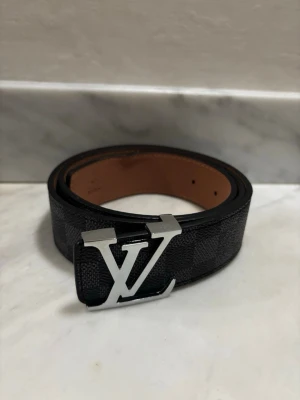 Louis Vuitton svart skinnbälte - Säljer ett svart skinnbälte från Louis Vuitton med klassiskt rutigt mönster och en silverfärgad LV-logga som spänne. Bältet har en lyxig känsla och är perfekt för att lyfta din outfit. Längd 105 cm.