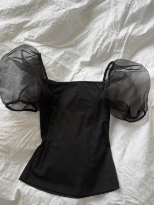 Svart topp med puffärmar - Trendig svart topp med stora puffärmar i transparent material. Toppen har en rak halsringning och är figurnära i modellen. Perfekt för dig som vill sticka ut med en cool och modern look.