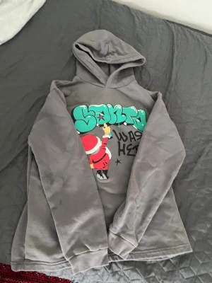 Grå hoodie med graffiti och tomte - Cool grå hoodie med huva och ett färgglatt graffiti-tryck på bröstet där en tomte sprayar texten 'Santa was here'. Långärmad och tillverkad i mjukt material, perfekt för dig som gillar streetstyle och unika prints.