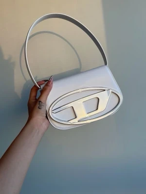 Vit handväska - Liten vit axelväska i stilren design. Slät yta med dekorativ detalj framtill och kort axelrem. Smidig storlek som rymmer det viktigaste. Passar både till vardag och kväll. 