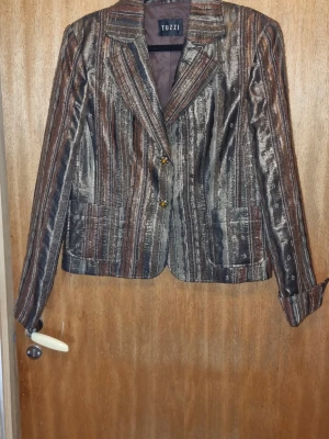 Randig kavaj från Tuzzi med smyckesknappar - Unik kavaj från Tuzzi med vertikala ränder i brunt, svart och guld. Kavajen har två framfickor och dekorativa smyckesknappar i guldfärg. Materialet har en glansig finish som ger en lyxig känsla. Perfekt för dig som vill sticka ut med din stil.