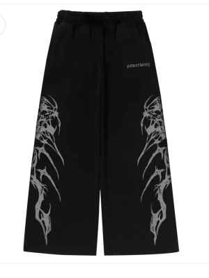 Kazuro pants - Svarta wide-leg byxor från Powerwing med elastisk midja och snörning. Byxorna har gråa tribal-liknande tryck längs benen och är tillverkade i ett syntetmaterial som ger en sportig känsla. Perfekta för dig som gillar streetwear och unika detaljer. Orginal pris är 520 kr