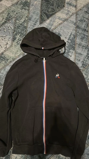 svart frankrike zip up hoodie - Svart hoodie med dragkedja från Le Coq Sportif. Har huva med snörning, fickor framtill och dragkedja i blått, vitt och rött. Broderad logga på bröstet. Perfekt för en sportig och avslappnad stil. passar både storlek XS och S