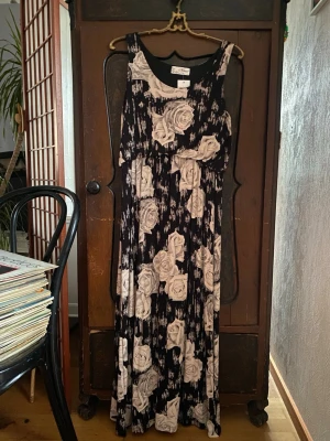 Svart långklänning med stora rosor - Långklänning i svart med stora vita rosor som mönster. Klänningen är ärmlös med rund halsringning och har en mjuk, följsam passform. Perfekt för dig som gillar blommiga prints och en stilren look.