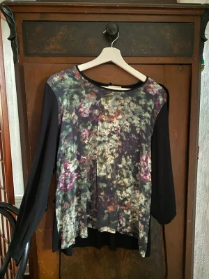 Blommig långärmad topp med svarta ärmar - Snygg långärmad topp med svart bas och blommigt mönster i grönt, lila och vitt framtill. Rund halsringning och lös passform. Perfekt för dig som gillar en unik och färgstark stil. (Möts upp i Stockholm)