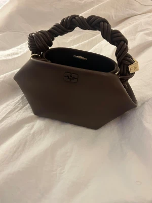 Ganni väska  - Säljer denna fina väska från ganni, sparsam använd. Mycket bra skick, ingen defekt på väskan. Köpt i Ganni butik i Köpenhamn! Väskan är i röd/brun färg och denna färg finns ej längre o köpa. Köpte denna väska maj 2025! Så vädligt ny ❤️Dustbag kommer med plus bandet som kommer med väskan! 