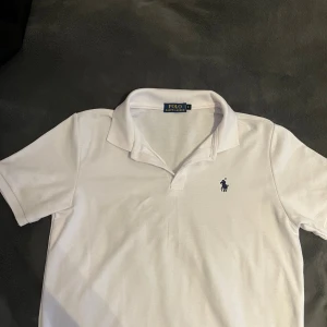 Vit pikétröja från Polo Ralph Lauren - Klassisk vit pikétröja från Polo Ralph Lauren med blå broderad logga på bröstet. Tröjan har korta ärmar, krage och knappar vid halsen. Perfekt för en stilren och sportig look.