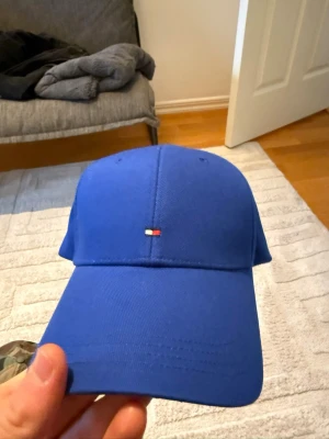 Blå keps från Tommy Hilfiger - Snygg blå keps från Tommy Hilfiger med det klassiska lilla flagglogotypen broderad framtill. Kepsen har böjd skärm och justerbart spänne bak. Tillverkad i bomull för en bekväm och stilren look.