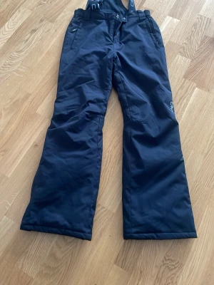 Svarta täckbyxor med hängslen från CMP - Säljer ett par svarta täckbyxor från CMP med justerbara hängslen och hög midja. Byxorna har raka ben, dragkedja och knapp framtill samt praktiska fickor. Perfekta för kalla dagar i backen eller snölek.