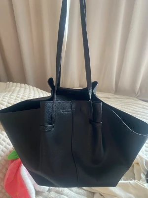 Svart shoppingväska i skinnimitation - Stilren svart shoppingväska med lång axelrem och mjuk struktur. Väskan har en enkel design utan synliga loggor och är tillverkad i skinnimitation med lätt kornig yta. Perfekt för dig som vill ha plats för allt och gillar minimalistisk look.