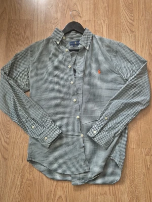 Rutig skjorta från Polo Ralph Lauren - Klassisk långärmad skjorta från Polo Ralph Lauren i grönvitt rutigt mönster. Skjortan har knappar framtill, button-down krage och en liten röd broderad logga på bröstet. Perfekt för en stilren och avslappnad look.