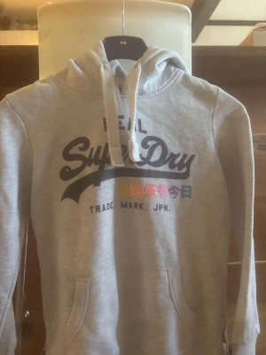 Ljusgrå hoodie från Superdry - Snygg ljusgrå hoodie från Superdry med stor logga och japansk text framtill. Hoodien har en klassisk huva med vita dragsnören och en känguruficka. Tillverkad i mjukt material som passar perfekt för en avslappnad stil.