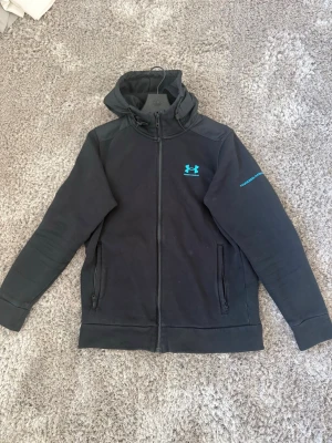 Under Armour - Snygg svart hoodie från Under Armour med dragkedja och huva. Har turkost logotryck på bröstet och text på ärmen. Tillverkad i mjukt material med förstärkta axelpartier och två fickor framtill. Perfekt för en sportig och avslappnad stil.