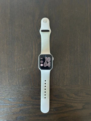 Apple Watch SE - Apple Watch smartklocka med rektangulär skärm och ljus sportrem. Klockan levereras med originalförpackning, laddkabel och laddplatta. I annonsen ingår även ett extra metallarmband i guldton som kan bytas mot sportremmen. Passar för vardagsanvändning, träning och notifieringar från ansluten iPhone.  Modell:  - Apple Watch SE (GPS) (2nd Generation) - 40mm Case - Aluminium.