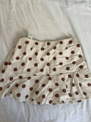 Vit kortkjol med bruna prickar - Söt kortkjol i vit bomull med stora bruna prickar och volang nedtill. Kjolen har en avslappnad passform och är perfekt för varma dagar. Enkel att matcha med både t-shirt och linne. Obs! Den hade tidigare inbyggda shorts, men dessa har jag tagit bort. Detta är däremot inget man märker på kjolen  