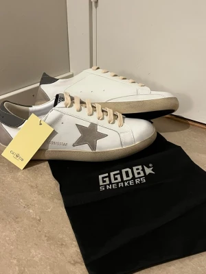 Golden Goose vita sneakers med stjärna - Snygga vita sneakers från Golden Goose med grå mockadetaljer, beige snören och ikonisk stjärna på sidan. Skorna har en låg silhuett och svart hälflik med logga. Kommer med originalpåse. Perfekta för en trendig streetstyle-look.