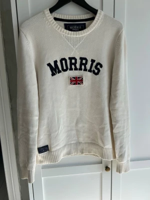 Vit stickad tröja från Morris - Vit stickad tröja från Morris med mörkblå text och en broderad brittisk flagga på bröstet. Tröjan har rund halsringning med ribbade muddar vid ärmslut och nederkant. Perfekt för dig som gillar klassisk och stilren design.