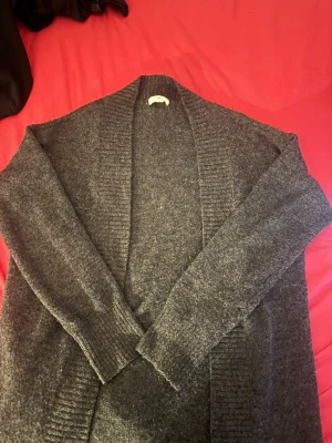 Mörkgrå lång kofta i mjukt material - Lång och mysig mörkgrå kofta med öppen front och bred ribbstickad kant. Koftan har långa ärmar och är gjord i ett mjukt, fluffigt material som känns skönt mot huden. Perfekt för lager på lager och chill dagar.