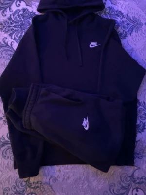Svart Nike hoodie med logga - Säljer en svart hoodie och byxorna  från Nike med klassisk vit logga på bröstet. Hoodien har huva med dragsko och ribbade muddar. Perfekt för en avslappnad och sportig stil. Materialet känns mjukt och bekvämt, och passformen är loose fit.