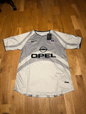Psg 01/02 away kit Ronaldinho #21 - Säljer en grå PSG bortatröja från Nike med Ronaldinho och nummer 21 på ryggen. Tröjan har svart Nike-logga, PSG-märke på bröstet och Opel som sponsor framtill. Tillverkad i lätt och ventilerande material, perfekt för fotboll eller samling.