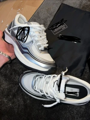 Chunky vita och silver sneakers - Säljer ett par chunky sneakers från  i vitt och silver med svarta detaljer. Skorna har en futuristisk design med mesh och läderpaneler, tjock sula och vita skosnören. Perfekta för dig som vill sticka ut med en lyxig och modern look.