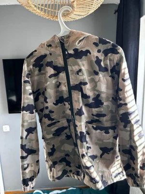 Camo Tracksuit - Säljer en camouflagemönstrad jumpsuit med huva och dragkedja framtill. Jumpsuiten har flera praktiska fickor, elastisk midja och muddar vid bensluten. Perfekt för dig som gillar streetwear och vill sticka ut med en edgy look. Storlek S. Fraktas timmen de köps. 