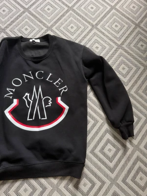 Svart sweatshirt från Moncler - Snygg svart sweatshirt från Moncler med stort vitt och rött logotryck på bröstet. Tröjan har rund halsringning, långa ärmar och ribbade muddar vid ärmslut och nederkant. Perfekt för en trendig och avslappnad stil.