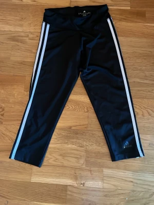 Svarta Adidas leggings med vita ränder - Säljer ett par svarta Adidas leggings i storlek XS med klassiska vita ränder längs sidorna. Tillverkade i climalite-material som andas och håller dig torr. Perfekta för träning eller chill. Adidas-logga på benet och hög midja för skön passform.
