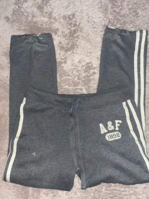 Mörkgrå mjukisbyxor från A&F - Mörkgrå mjukisbyxor från Abercrombie & Fitch med vita ränder längs sidorna och broderad logga 'A&F 1892' på vänster ben. Storlek L men sitter som M