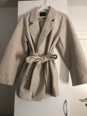 Lager157 beige kappa med knytskärp, strl M - Stilren beige kappa från Lager157 med knytskärp i midjan. Kappan har raka ärmar, två fickor framtill och är tillverkad i polyester material. Perfekt för dig som gillar minimalistisk och trendig stil. Tyvärr har den några fläckar, en hål oxh noppor som syns på bilderna. Jag har  försökt göra rent och få bort nopporna, men det finns fortfarande få kvar. Så ifall man har en noppor-borttagare så funkar det bäst! 