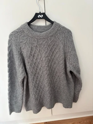 Grå kabelstickad tröja - Mysig grå kabelstickad tröja med rund halsringning och ribbade muddar. Tröjan har ett klassiskt flätmönster över hela framsidan och är tillverkad i ett mjukt, tjockt material som passar perfekt för kyliga dagar. Långärmad och oversized passform. Strl M. Något nopprig, tecken på användning. Inga hål/fläckar 💛