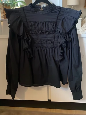 Svart volangblus med hög krage - Stilren svart blus med hög krage och dekorativa volanger framtill och på axlarna. Blusen har långa ärmar och fina detaljer som rynk och små bollar längs kanterna. Perfekt för dig som gillar en trendig och romantisk look. I storlek M, använt ett enstaka gånger men är för kort i armarna för mig. Pris kan diskuteras❤️