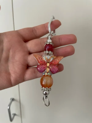Nyckelring med fjäril och pärlor - Säljer en färgglad nyckelring med en dekorativ fjäril i orange och rosa nyanser, glittriga pärlor och silvriga detaljer. Perfekt att hänga på väskan eller nyckelknippan för en personlig touch.