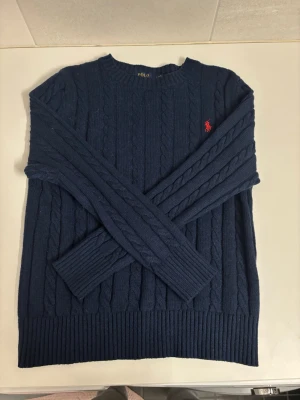 Mörkblå kabelstickad tröja Polo Ralph Lauren - Kabelstickad mörkblå tröja från Polo Ralph Lauren med rund halsringning och klassisk röd broderad logga på bröstet. Tröjan har ribbade muddar vid ärmslut och nederkant. Perfekt för dig som gillar stilrena och tidlösa plagg.