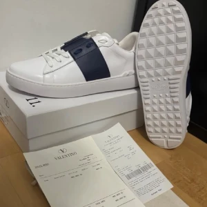 Nya Valentino vita sneakers - Snygga vita sneakers från Valentino med en bred rem över sidan och matchande blå detaljer vid snörningen. Skorna har en klassisk låg profil, vit sula med rutmönster undertill och är tillverkade i slätt läder. Perfekta för en clean och stilren look.